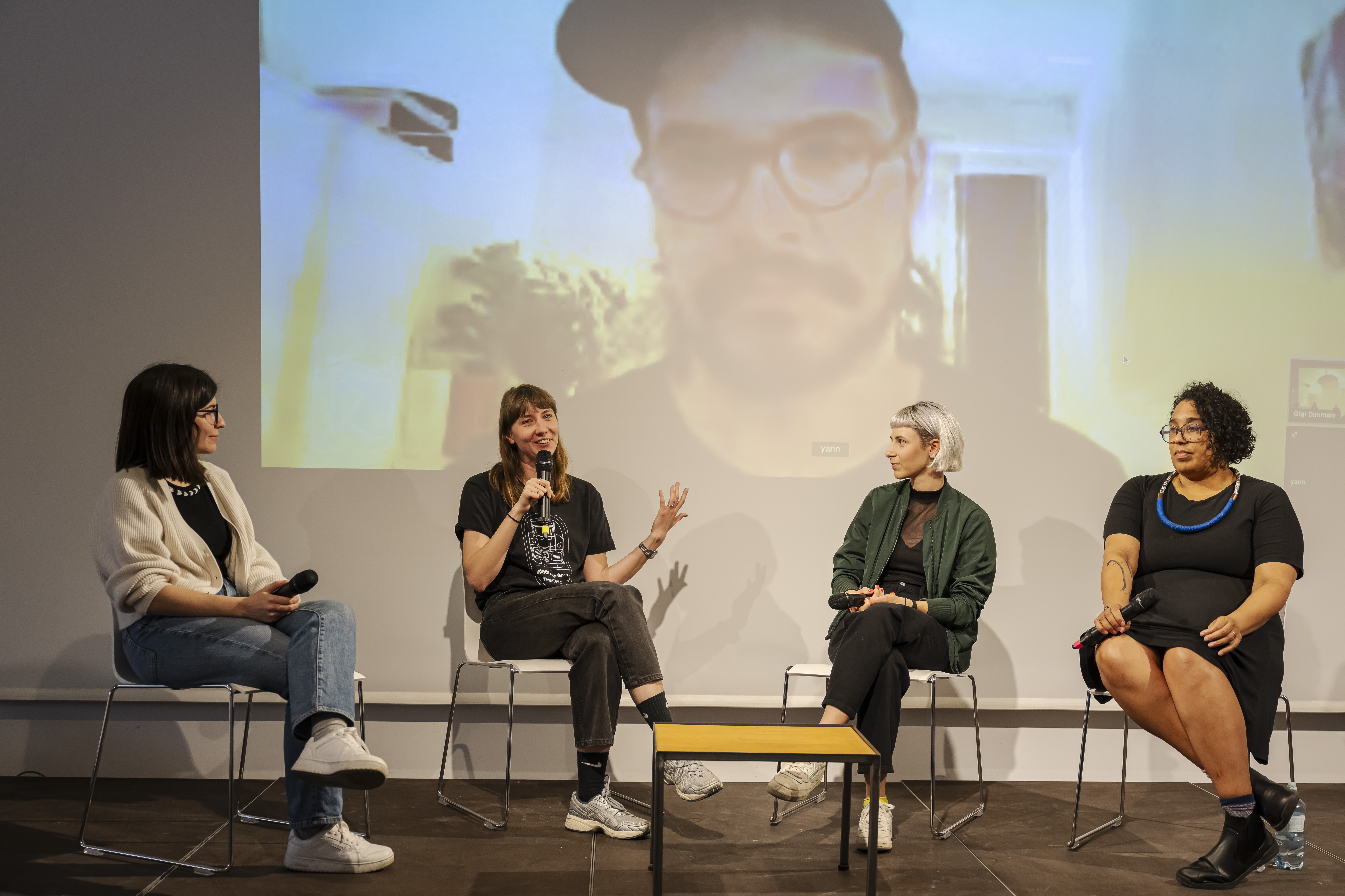 four people sitting on a stage on chairs talking. behind their back, projection with the video from the fifth participant
